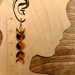 May include: A pair of brown and gold moon phase earrings. The earrings are made of resin and have a dangling design. The earrings are about 4 inches long.