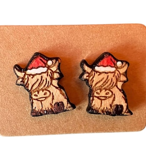 May include: A pair of wooden stud earrings featuring a Highland cow design. Each cow is wearing a red Santa hat with white trim. The earrings are light brown with dark brown and red accents, set on a brown card.