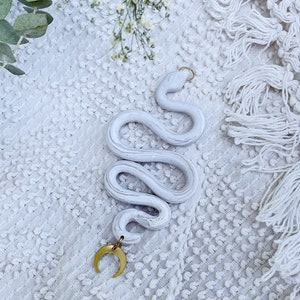 May include: White ceramic snake figurine with a gold crescent moon charm. The snake is curled up in a coil shape and has a textured surface. The figurine is on a white textured background.