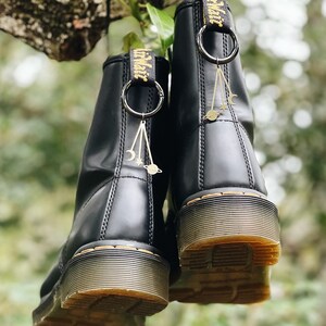 Puede incluir: Un par de botas de cuero negro con una etiqueta amarilla que dice "Dr. Martens" colgando de la parte superior de cada bota. Cada bota tiene un amuleto dorado colgando de un anillo plateado en la etiqueta. Los amuletos son una luna creciente, una estrella y un planeta.