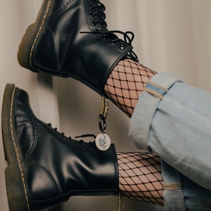 Könnte beinhalten: Schwarze Lederstiefel mit Schnürung, gelben Nähten und einem gelben und schwarzen Anhänger, der an den Schnürsenkeln befestigt ist. Die Stiefel werden mit Netzstrümpfen und einer blauen Jeans getragen.
