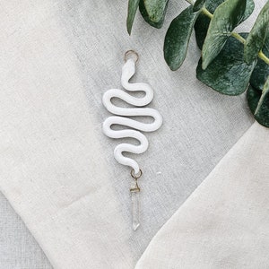 May include: White ceramic snake charm with a clear quartz crystal pendant. The charm is hanging from a gold chain.