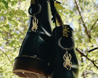 May include: A pair of black leather boots with yellow stitching and gold beetle charms hanging from the laces. The boots are hanging from a tree branch.