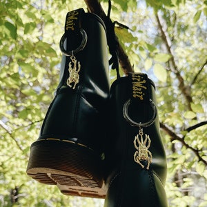 Może przedstawiać: Para czarnych skórzanych butów z żółtymi szwami i złotymi zawieszkami w kształcie chrząszcza wiszącymi na sznurowadłach. Buty wiszą na gałęzi drzewa.