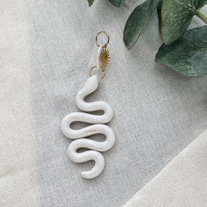 May include: White ceramic snake charm pendant with a gold-toned clasp. The pendant is hanging from a white fabric background.