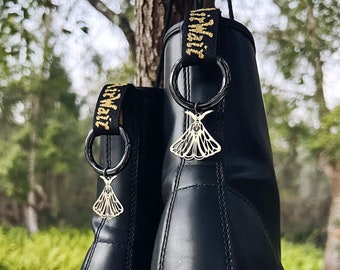May include: A pair of black leather boots with gold accents and a gold moth charm hanging from each boot. The boots are hanging from a tree branch.