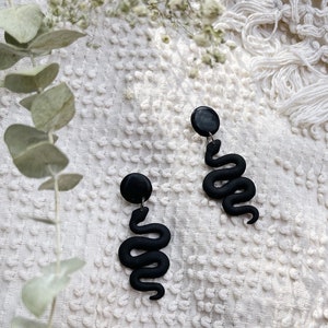 May include: A pair of black snake-shaped dangle earrings. The earrings are made of polymer clay and have a black disc at the top.