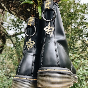 Op de afbeelding: Een paar zwarte leren laarzen met een gele slangenhanger aan elke laars. De laarzen hebben een dikke, zwarte zool en een gele stikdetail rond de rand van de zool.