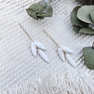 May include: A pair of white ceramic earrings with a gold wire design. The earrings are shaped like leaves and have a speckled texture.