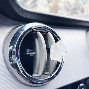 May include: A close-up of a car's air vent with a clear crystal hanging from it. The vent is made of chrome and has a black background. The crystal is hanging from a small hook.