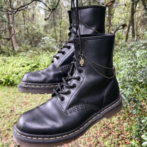 May include: A pair of black leather Dr. Martens boots with yellow stitching and laces. The boots are hanging from a tree branch with a gold chain and a cactus charm.