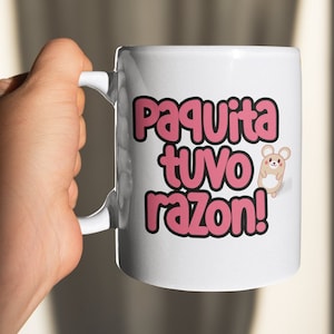 May include: White ceramic mug with the text "Paquita tuvo razon!" in pink bubble letters and a cartoon mouse illustration. The mug is held by a hand, with a plain background.