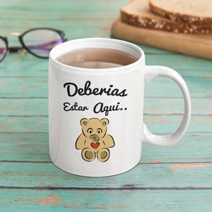 May include: White ceramic mug with the text "Deberias Estar Aqui.." and a cartoon teddy bear with a red heart. The mug is filled with a brown liquid and sits on a teal wooden surface. A pair of glasses and bread slices are in the background.