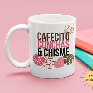 Może przedstawiać: Biały ceramiczny kubek z napisem "CAFECITO CONCHAS & CHISME" w brązowych i r&oacute;żowych literach. Kubek przedstawia ilustracje conchas, rodzaju meksykańskiego słodkiego chleba. R&oacute;żowy i czerwony notatnik znajduje się w tle.