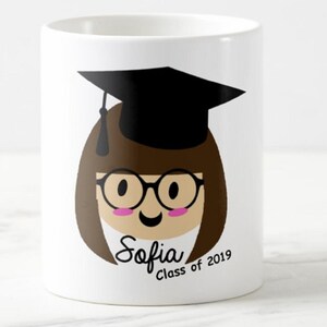 May include: White ceramic mug featuring a cartoon illustration of a graduate with brown hair, glasses, and a black graduation cap. The mug has the name "Sofia" and "Class of 2019" printed on it. A celebratory gift for a graduate.