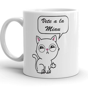 May include: White ceramic mug featuring a cartoon cat with a speech bubble. The cat is drawn in black outline with pink inner ears and a sad expression. The speech bubble says "Vete a la Miau" in black script.