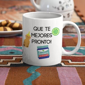 Może przedstawiać: Biały ceramiczny kubek z napisem "QUE TE MEJORES PRONTO!" oraz grafiką z kresk&oacute;wkowym misiem, plasterkiem limonki i butelką z lekarstwem. Biały imbryk i ciastka w tle.