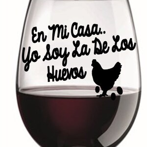 May include: A wine glass filled with red wine. The glass has black text that reads "En Mi Casa... Yo Soy La De Los Huevos" with a silhouette of a hen and eggs. The glass is on a white background.