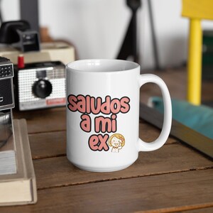 Może przedstawiać: Biały ceramiczny kubek z napisem "saludos a mi ex" w r&oacute;żowych i czarnych literach. Obok tekstu znajduje się rysunkowy obraz uśmiechniętej kobiety. Kubek stoi na drewnianej powierzchni z zabytkowymi aparatami w tle.