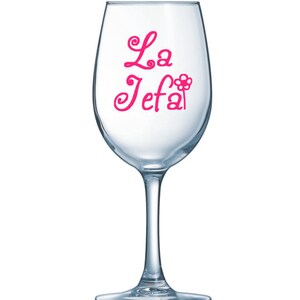 May include: A clear wine glass with a long stem. The words "La Jefa" are written in pink cursive script on the glass, with a flower graphic replacing the "a" in "Jefa".