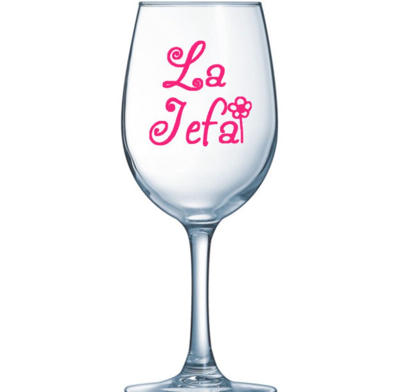 May include: A clear wine glass with a long stem. The words "La Jefa" are written in pink cursive script on the glass, with a flower graphic replacing the "a" in "Jefa".