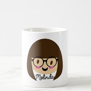 May include: White ceramic mug featuring a cartoon illustration of a person with brown hair, glasses, and a smiling face. The name "Melinda" is written in cursive below the face. The mug is cylindrical and has a glossy finish.
