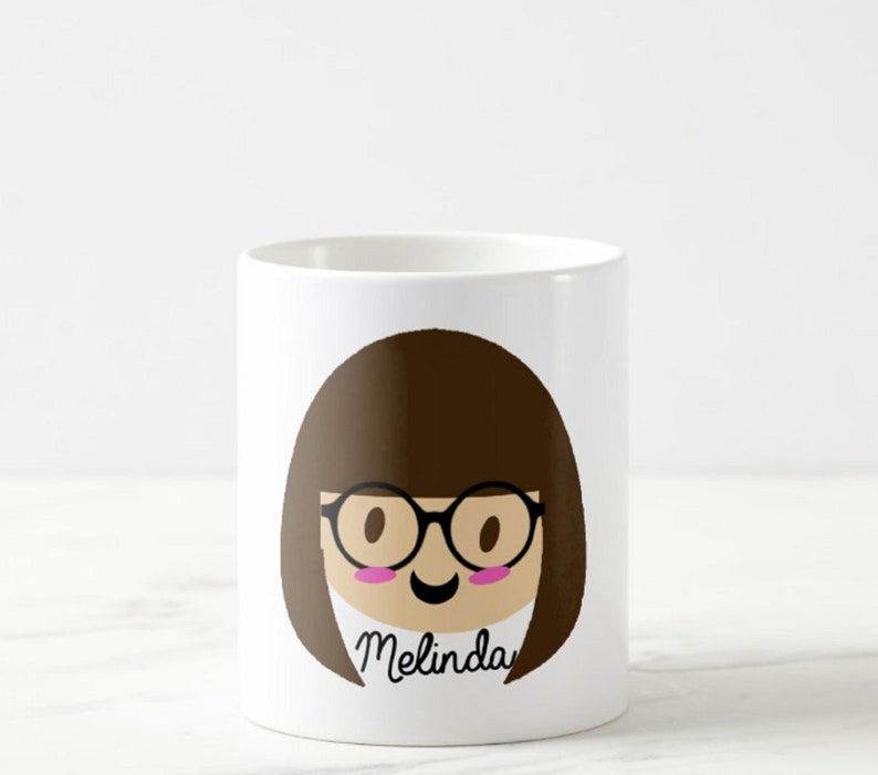 May include: White ceramic mug featuring a cartoon illustration of a person with brown hair, glasses, and a smiling face. The name "Melinda" is written in cursive below the face. The mug is cylindrical and has a glossy finish.