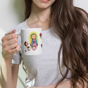 May include: White ceramic mug featuring a colorful illustration of a religious figure with a halo, surrounded by cartoon figures and red roses. The mug has a curved handle and is held by a person wearing a gray t-shirt.
