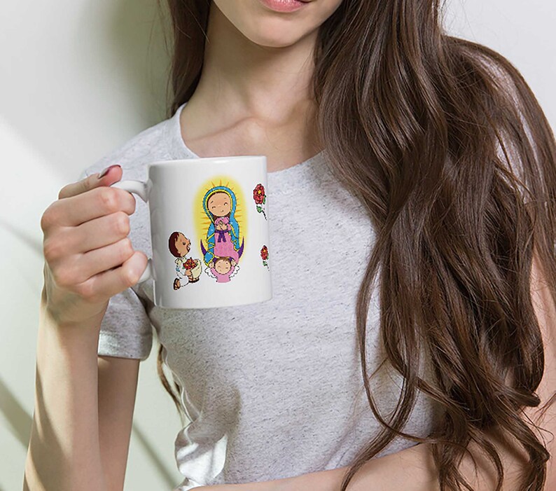 May include: White ceramic mug featuring a colorful illustration of a religious figure with a halo, surrounded by cartoon figures and red roses. The mug has a curved handle and is held by a person wearing a gray t-shirt.