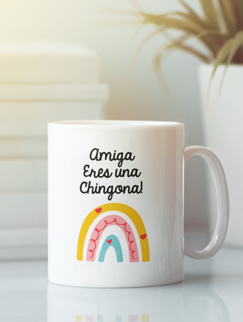 以下が含まれることがあります： 「Amiga Eres una Chingona!」の文字と虹のデザインが施された白いセラミックマグ。虹は、黄色、ピンク、赤、青のアーチとハートのアクセントが特徴です。マグカップには湾曲したハンドルが付いています。