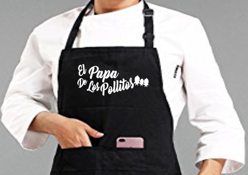 Pu&ograve; includere: Grembiule nero con la scritta bianca "El Papa De Los Pollitos". Il grembiule ha una tasca e nella tasca &egrave; visibile un telefono rosa. Il grembiule &egrave; indossato sopra una giacca da chef bianca.