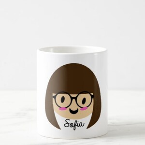 May include: White ceramic mug featuring a cartoon illustration of a person with brown hair, glasses, and a smiling face. The name "Sofia" is written in black script below the face. The mug is cylindrical.