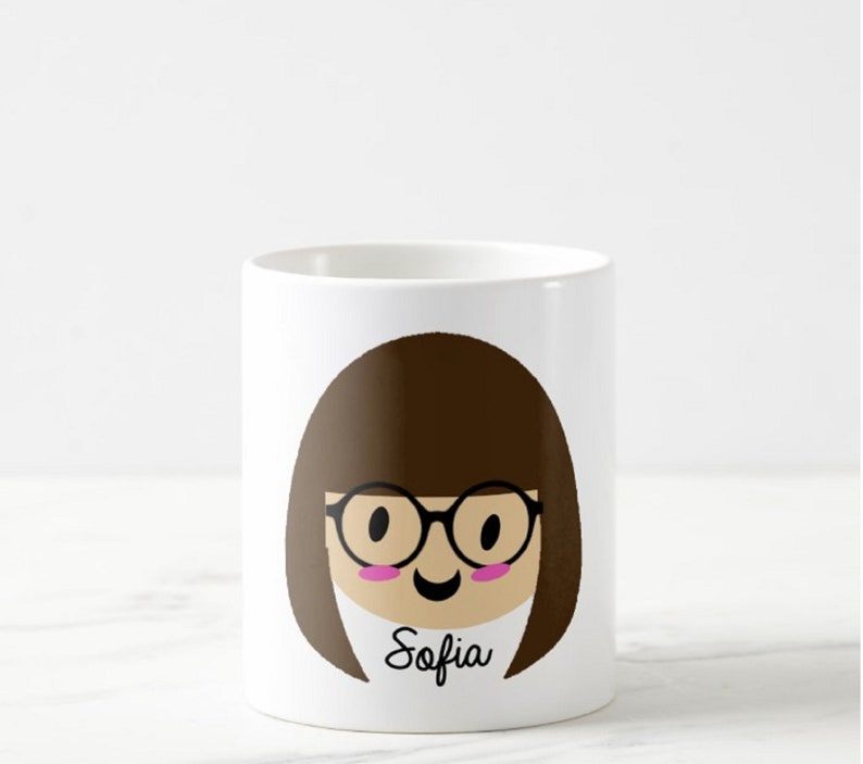 May include: White ceramic mug featuring a cartoon illustration of a person with brown hair, glasses, and a smiling face. The name "Sofia" is written in black script below the face. The mug is cylindrical.