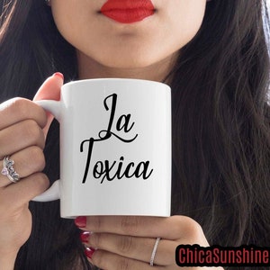 以下が含まれることがあります： 黒い筆記体で「La Toxica」と書かれた白いセラミックマグ。赤い口紅と黒髪の人がマグカップを持っています。ChicaSunshine.comのウェブサイトが画像に表示されています。