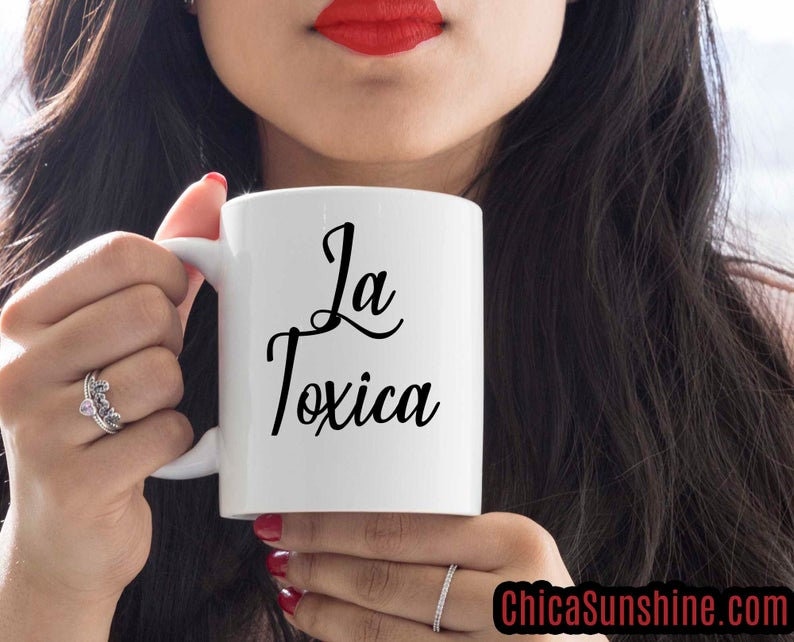 以下が含まれることがあります： 黒い筆記体で「La Toxica」と書かれた白いセラミックマグ。赤い口紅と黒髪の人がマグカップを持っています。ChicaSunshine.comのウェブサイトが画像に表示されています。