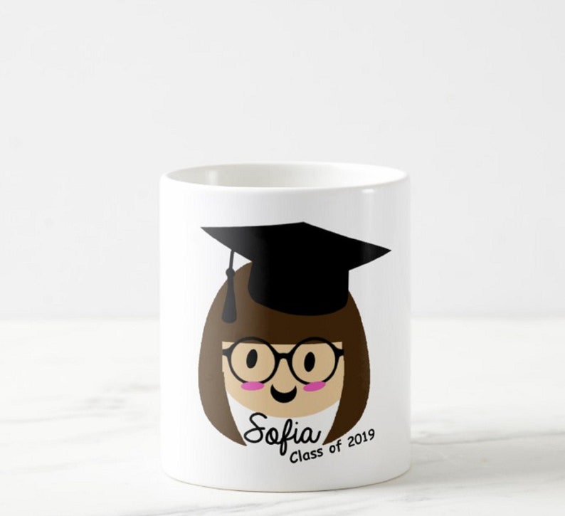 May include: White ceramic mug featuring a cartoon illustration of a graduate with brown hair, glasses, and a black graduation cap. The mug has the name "Sofia" and "Class of 2019" printed on it. A celebratory gift for a graduate.