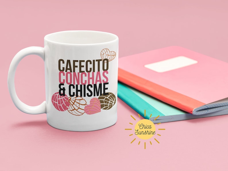 Może przedstawiać: Biały ceramiczny kubek z napisem "CAFECITO CONCHAS & CHISME" w brązowych i r&oacute;żowych literach. Kubek przedstawia ilustracje conchas, rodzaju meksykańskiego słodkiego chleba. R&oacute;żowy i czerwony notatnik znajduje się w tle.