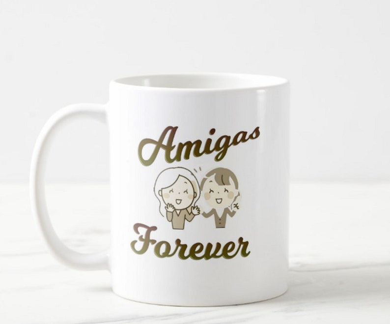 Może przedstawiać: Biały ceramiczny kubek z napisem "Amigas Forever" w złotym skrypcie. Kubek przedstawia kresk&oacute;wkową ilustrację dw&oacute;ch uśmiechniętych postaci. Kubek ma zakrzywiony uchwyt i nadaje się do gorących lub zimnych napoj&oacute;w.