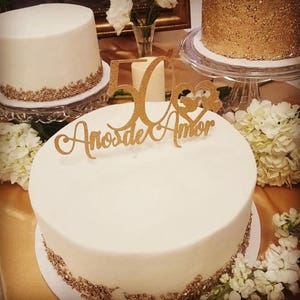 Puede incluir: Varios pasteles glaseados blancos con detalles dorados y un adorno dorado que dice "50 A&ntilde;os de Amor". Los pasteles est&aacute;n sobre soportes de vidrio y rodeados de flores blancas. Un pastel tiene un acabado dorado con lentejuelas.