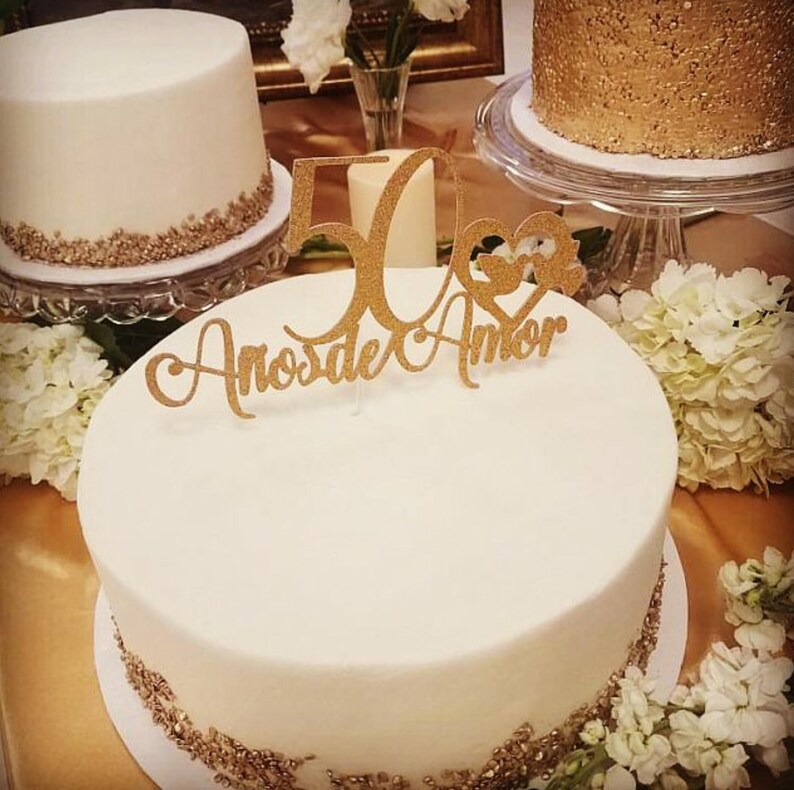 Puede incluir: Varios pasteles glaseados blancos con detalles dorados y un adorno dorado que dice "50 A&ntilde;os de Amor". Los pasteles est&aacute;n sobre soportes de vidrio y rodeados de flores blancas. Un pastel tiene un acabado dorado con lentejuelas.