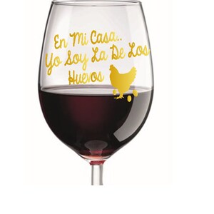 May include: A clear wine glass filled with red wine. The glass has gold lettering and a gold chicken graphic. The text reads "En Mi Casa... Yo soy La De Los Huevos".