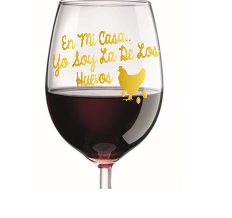 May include: A clear wine glass filled with red wine. The glass has gold lettering and a gold chicken graphic. The text reads "En Mi Casa... Yo soy La De Los Huevos".
