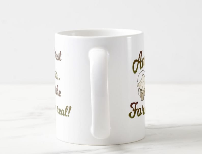 Może przedstawiać: Biały ceramiczny kubek do kawy z zakrzywionym uchwytem. Kubek ma złoty napis &bdquo;I pierwszy, kt&oacute;ry... zobaczy prawdziwe!&rdquo; oraz rysunkową ilustrację kobiety.