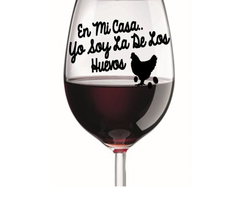 May include: A wine glass filled with red wine. The glass has black text that reads "En Mi Casa... Yo Soy La De Los Huevos" with a silhouette of a hen and eggs. The glass is on a white background.
