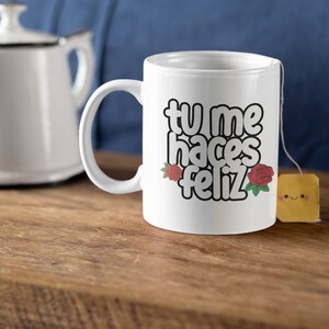 May include: White ceramic mug with the Spanish text "tu me haces feliz" in a black outline font, decorated with red roses. A tea bag with a smiling face is attached to the mug. A white teapot is in the background.