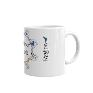 May include: White ceramic mug with a handle, featuring the name "Regina" in black script and a blue butterfly. The mug has a floral design in shades of blue, purple, and gold. Text in Spanish is also visible.