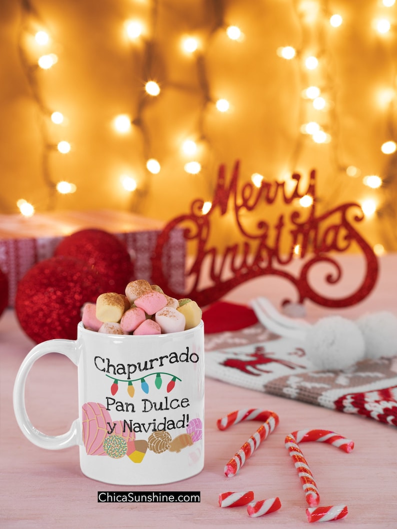 May include: A white mug filled with marshmallows, featuring the text "Chapurrado Pan Dulce y Navidad!" and colorful illustrations of pastries. Candy canes and Christmas decorations are scattered around the mug, with a blurred background of Christmas lights.