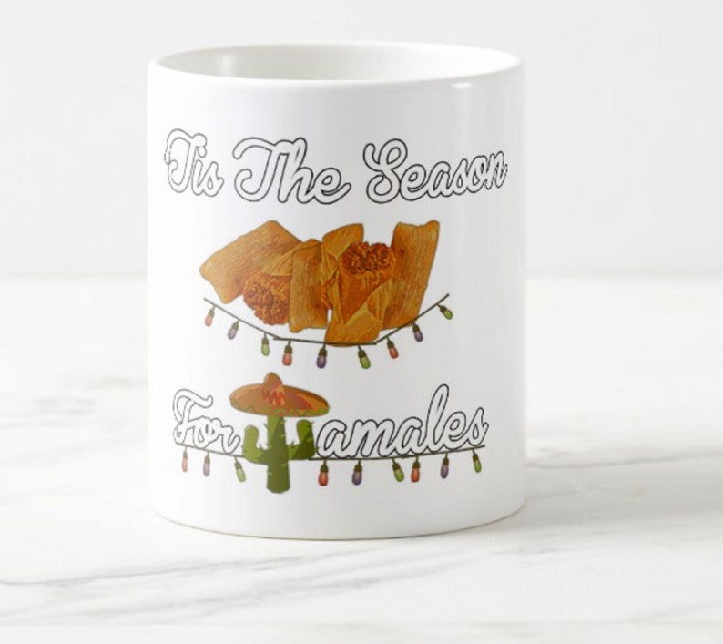 以下が含まれることがあります： 「Tis The Season For Tamales」の文字が入った白いセラミックマグ。マグには、タマレス、ソンブレロ、サボテンのグラフィックが描かれており、すべてストリングライトで飾られています。