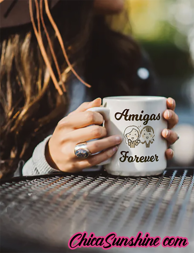 以下が含まれることがあります： 「Amigas Forever」の文字が黒い筆記体で書かれた白いセラミックマグ。2人の友人の漫画イラストが描かれています。マグカップは指輪をつけた人が持っています。画像には「ChicaSunshine com」の文字も含まれています。