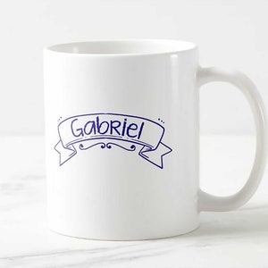 Puede incluir: Taza de cer&aacute;mica blanca con el nombre "Gabriel" en azul dentro de un dise&ntilde;o de banner. La taza tiene un asa curva y un dise&ntilde;o simple y limpio. El texto es de estilo manuscrito.